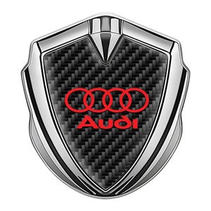 May include: A silver and black car emblem with a red Audi logo. The emblem has a carbon fiber pattern and is shaped like a shield.