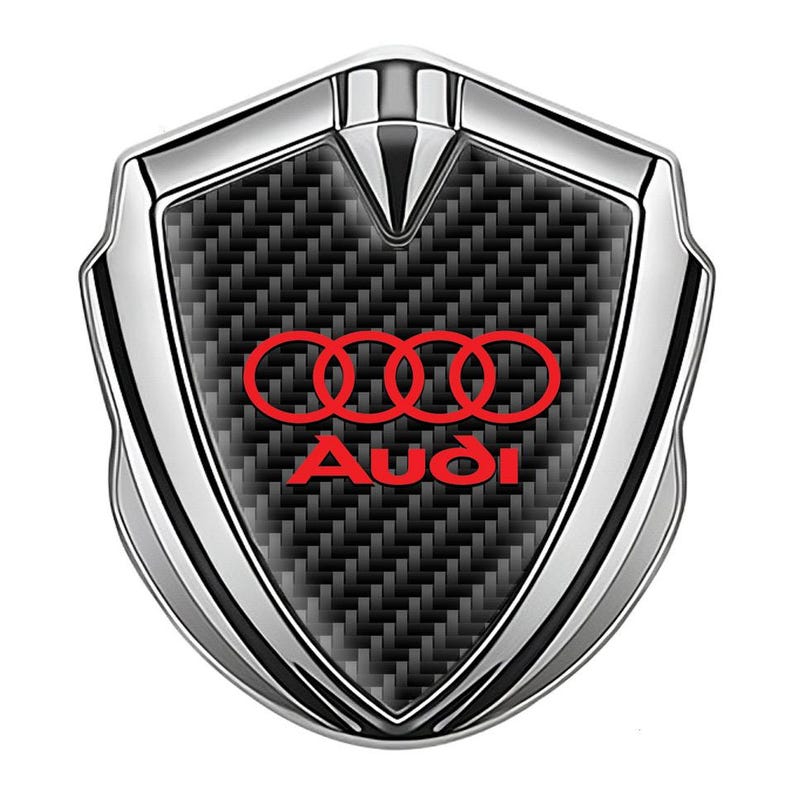 May include: A silver and black car emblem with a red Audi logo. The emblem has a carbon fiber pattern and is shaped like a shield.