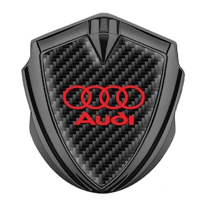 Könnte beinhalten: Ein schwarzes, schildförmiges Emblem mit einem Karbonfaser-Muster und einem roten Audi-Logo. Das Logo zeigt vier ineinandergreifende Ringe und das Wort "Audi" in Rot.