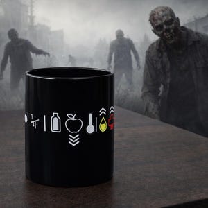 Op de afbeelding: Zwarte keramische mok met witte grafische iconen, waaronder een appel, fles en lepel. De mok staat op een houten oppervlak met een vage achtergrond van zombies in een mistige, post-apocalyptische setting.