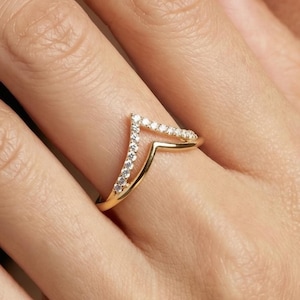 Könnte beinhalten: Ein goldfarbener Ring mit V-förmigem Design. Der Ring ist mit kleinen, klaren Edelsteinen besetzt. Der Ring befindet sich an einem Finger, mit Nahaufnahme von Hand und Gesicht. Modernes, elegantes Design.