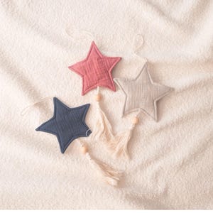 May include: Three fabric star ornaments with tassels hanging from a white surface. The stars are pink, gray, and blue.