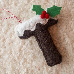 Può includere: Un ornamento fatto a mano a forma di budino di Natale. Il budino di feltro marrone è sormontato da 'glassa' di feltro bianco, foglie di agrifoglio di feltro verde e bacche di pompon rosse. Un anello di corda rosso e bianco è attaccato.