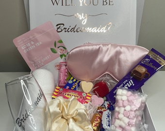 Può includere: Scatola regalo bianca con testo dorato "WILL YOU BE my Bridesmaid?". Contiene una mascherina per dormire rosa, una maschera per il viso, un calice da champagne, dolci e una barretta di cioccolato. Un nastro bianco avvolge la scatola.
