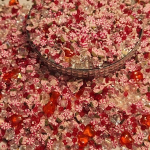 Pode incluir: Um close-up de uma variedade de confeitos coloridos, incluindo bast&otilde;es de caramelo rosa e branco, cora&ccedil;&otilde;es vermelhos e laranja, e strass transparentes e vermelhos. Os confeitos est&atilde;o em uma tigela e est&atilde;o prontos para serem usados para decorar bolos, cupcakes ou outras sobremesas.