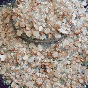 May include: A large pile of white, brown, and iridescent glitter with various shapes, including hearts, stars, and doughnuts. The glitter is in a clear glass bowl.
