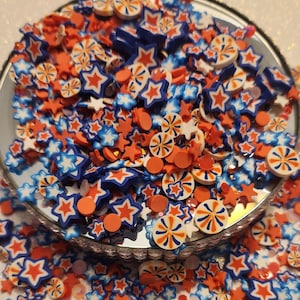 May include: A close-up of a bowl filled with red, white, and blue polymer clay sprinkles. The sprinkles are shaped like stars and swirls.
