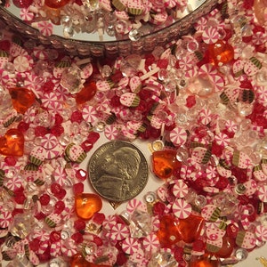 Pode incluir: Um close-up de uma variedade de confeitos rosa, branco e laranja, doces e contas. Os confeitos t&ecirc;m a forma de cora&ccedil;&otilde;es, estrelas e redemoinhos. H&aacute; uma moeda de um quarto de d&oacute;lar americano na imagem para dar uma ideia de escala.