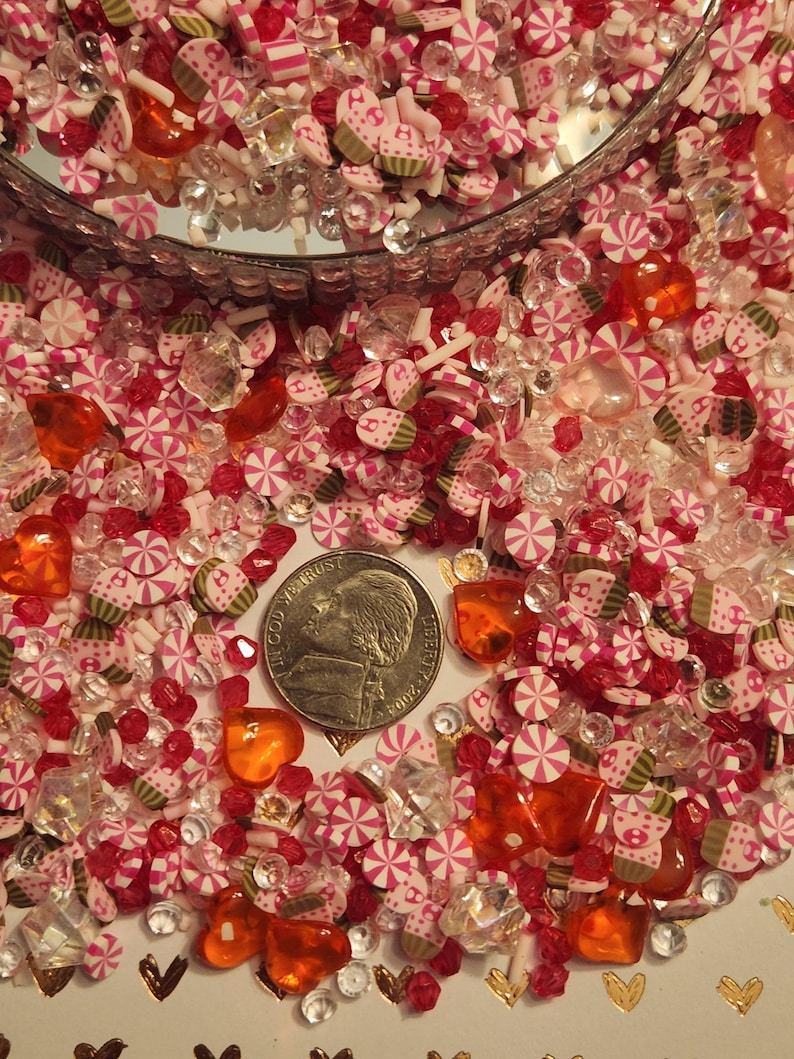 Pode incluir: Um close-up de uma variedade de confeitos rosa, branco e laranja, doces e contas. Os confeitos t&ecirc;m a forma de cora&ccedil;&otilde;es, estrelas e redemoinhos. H&aacute; uma moeda de um quarto de d&oacute;lar americano na imagem para dar uma ideia de escala.