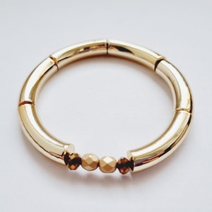 Peut inclure: Un bracelet doré composé d'une série de tubes ronds reliés. Le bracelet comporte trois perles dorées au centre.
