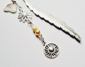 Op de afbeelding: Een zilverkleurige metalen bladwijzer met een lange veerhanger. Een bungelende ketting bevat een kleine regenboog-emaille bedel, een zilveren bloemvorm, twee lichtgele kralen en een ronde zonvormige bedel aan het uiteinde.