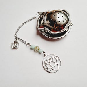 Pode incluir: Infusor de chá prateado com corrente e berloques decorativos. O infusor tem um corpo redondo e perfurado e uma tampa. A corrente é adornada com contas e um pingente de flor de lótus. Projetado para infusão de chá a granel.