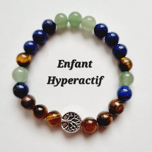Puede incluir: Una pulsera de cuentas con piedras verdes, azules y marrones, y un dije de árbol de la vida plateado. La pulsera tiene las palabras "Enfant Hyperactif" en texto negro.