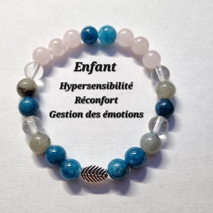 Puede incluir: Una pulsera de cuentas azules, rosas y transparentes, con un dije de hoja plateada. La pulsera tiene el texto "Enfant, Hypersensibilité, Réconfort, Gestion des émotions."
