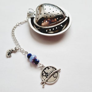 Könnte beinhalten: Silberner herzförmiger Tee-Infuser mit einer Kette und einem Anhänger. Der Anhänger zeigt einen Halbmond, drei blaue Perlen und einen runden Anhänger mit einem Stern und einer Katzensilhouette.