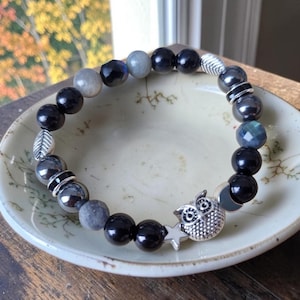 Peut inclure: Un bracelet de perles composé de pierres rondes noires, grises et bleu irisé, alternées avec des éléments en métal argenté. Le bracelet comporte une breloque hibou argentée, une petite étoile et deux intercalaires en forme de feuille.
