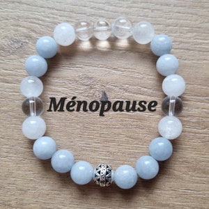 Peut inclure: Un bracelet en cristal bleu clair et transparent avec un charme argenté. Le bracelet porte le mot "Ménopause" écrit sur fond blanc.