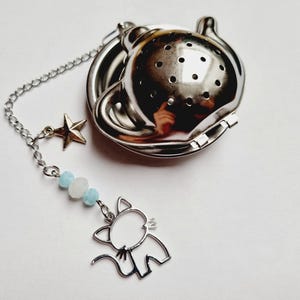 Peut inclure: Infuseur à thé en acier inoxydable avec un charm en forme de chat et un pendentif étoile. L'infuseur a une chaîne avec des perles bleues et blanches. Conçu pour le thé en vrac.