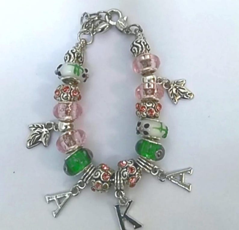 Pode incluir: Pulseira de berloques prateada com contas rosa, verdes e brancas. A pulseira apresenta berloques de folhas e berloques de letras que soletram "AKA". As contas t&ecirc;m uma variedade de texturas e designs, incluindo detalhes de strass.
