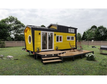 May include: A small yellow and black house with a wooden deck and steps. The house has a large window and a small round window. There is a grill on the grass.