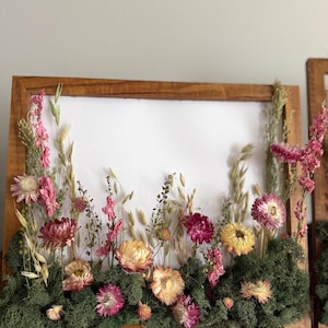 May include: A wooden framed artwork featuring a display of dried flowers and greenery. The arrangement includes pink, yellow, and burgundy blooms set against a backdrop of white paper. The base is covered in green moss.