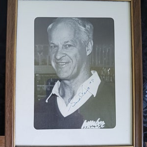 May include: A black and white photo of a man in a black sweater with a white collar. The man is smiling and has a blue signature that says "Sam Snead #9" and "member 1962" below the photo.