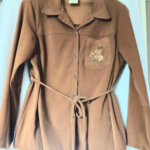 May include: A brown button-down shirt with a tie-waist and a small pocket on the chest. The pocket features an embroidered floral design. The shirt has long sleeves and a collared neckline. The fabric appears to be a soft, suede-like material.