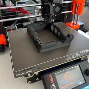 May include: A 3D printer with a black and orange frame is printing a black plastic object. The printer bed is gray and has the text "ORIGINAL PRUSA by Josef Prusa" printed on it. The printer screen shows a progress bar that is 56% complete.