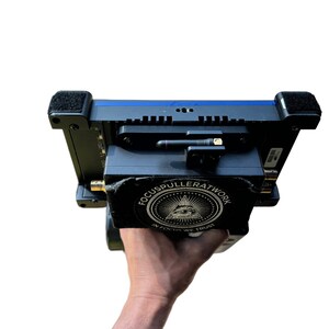 Puede incluir: Un dispositivo electrónico negro y azul con una pegatina que dice "FOCUSPULLERATWORK" y "IN FOCUS WE TRUST". El dispositivo tiene una pequeña antena y está sostenido por una mano.