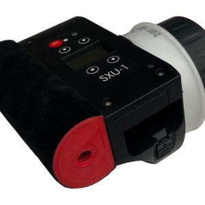 May include: Black and red handheld device with a white lens and a digital display that reads "SXU-1".