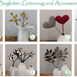 Op de afbeelding: Een verzameling gehaakte bloemstukken in verschillende kleuren en stijlen, tentoongesteld in witte keramische vazen. De arrangementen omvatten een ​​boeket kleine, paarse en witte bloemen, een takje gele bloemen, een paar rode en grijze harten, een paar groene bladeren, een boeket zwart-witte bloemen, een takje groene bladeren en een paar bruine bloemen.