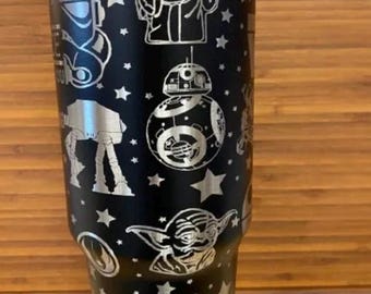 May include: A black stainless steel tumbler with a silver Star Wars themed design. The design features characters such as Yoda, R2-D2, and Stormtroopers. The text "May the Force Be With You" is printed on the bottom of the tumbler.