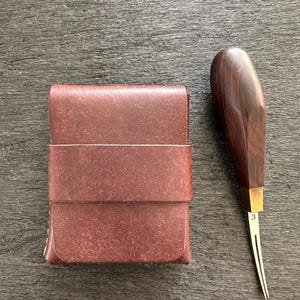 Puede incluir: Un tarjetero rectangular de cuero marrón oscuro con una correa horizontal en el centro y una herramienta de marcado de bordes de metal con mango de madera oscura y una virola de latón marcada con el número 3 situada a la derecha del objeto.