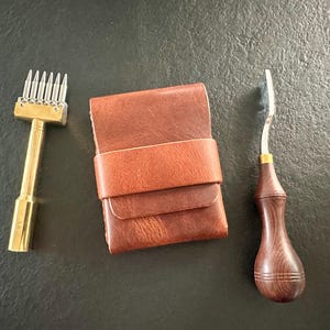 Puede incluir: Una cartera de cuero marrón con cierre de correa horizontal, situada entre un cincel de costura de seis puntas de latón y una herramienta biseladora de bordes de metal con mango de madera marrón oscuro, sobre una superficie texturizada.