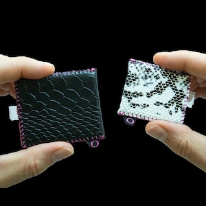 May include: Two small square pouches with a textured snakeskin pattern. One pouch is solid black, and the other is white with a black snakeskin print. Both feature bright pink stitched edges and a small pink circular tab on the bottom corner.