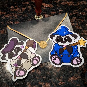 May include: Two panda bear stickers, one wearing a blue wizard hat and holding a gold wand, the other holding a paintbrush. Both stickers are on a gray envelope.
