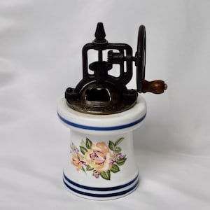 May include: Antique manual coffee grinder. The grinder features a white ceramic base with a floral design and blue stripes. The grinding mechanism is black metal with a wooden handle. A vintage kitchen decor item.