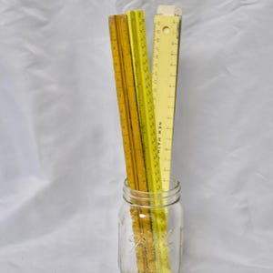 May include: A clear glass mason jar filled with three yellow rulers. The rulers are of varying shades of yellow and are standing upright. The background is a white, textured fabric.
