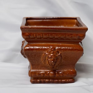 May include: A square, brown ceramic planter with a detailed, ornate design. The planter features relief carvings of faces and decorative trim. The top opening is square, and the overall style is classical.