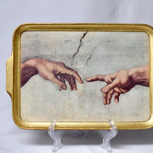 May include: A rectangular serving tray with a metallic gold frame and integrated handles. The center features a reproduction of the Creation of Adam fresco, depicting two reaching hands against a light, cracked, off-white plaster background.
