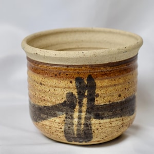 May include: A handmade ceramic pot with a beige rim and a textured, speckled surface. The pot features horizontal bands of brown and tan hues, with a dark brown abstract design. This decorative piece is suitable for home decor.