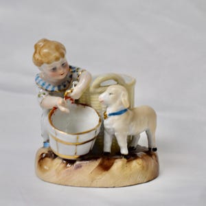 May include: A porcelain figurine depicting a child with a bucket, a lamb, and a basket. The child is dressed in a blue and white outfit. The lamb wears a blue collar. The base is tan and brown.