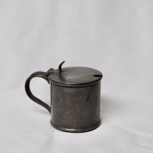 May include: A small, antique pewter mug with a lid and handle. The mug is cylindrical with a curved handle and a lid with a small, curved handle. The pewter has a dark, aged appearance. The mug is likely used for serving or storing liquids.