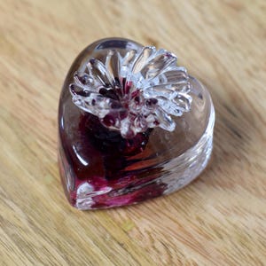 May include: A clear glass heart-shaped paperweight with a flower design on top. The heart is filled with a deep red color. The paperweight is sitting on a wooden surface.