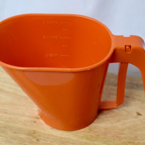 May include: Orange plastic measuring cup with a handle. The cup has a wide, oval opening and a tapered body. Measurement markings are visible on the interior. The handle is attached to the side of the cup.