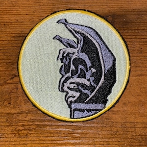 May include: Embroidered patch featuring a stylized gargoyle in shades of black and gray. The gargoyle is set against a light green background within a circular shape, outlined with a yellow border.