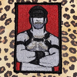 May include: Embroidered patch featuring a stylized portrait of a person with a beard and cap, wearing a harness. The portrait is set against a red background, framed in black, and placed on a leopard print background.