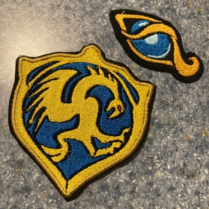 Könnte beinhalten: Zwei gestickte Aufnäher. Einer ist schildförmig mit einem goldenen Rand und einem blau-goldenen Design. Der andere ist ein augenförmiger Aufnäher mit einem blauen Auge und goldenen Akzenten. Beide Aufnäher haben schwarze Ränder.