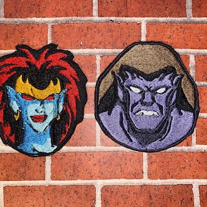 Könnte beinhalten: Zwei gestickte Aufnäher mit Cartoon-Wasserspeier-Charakteren. Ein Aufnäher zeigt einen weiblichen Wasserspeier mit blauer Haut, roten Augen und einer goldenen Krone. Der andere Aufnäher zeigt einen männlichen Wasserspeier mit violetter Haut. Beide haben schwarze Ränder.
