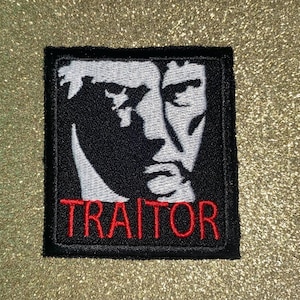 May include: Black and white embroidered patch featuring a stylized face. The word "TRAITOR" is embroidered in red letters at the bottom of the patch. The patch has a black border and is square in shape.